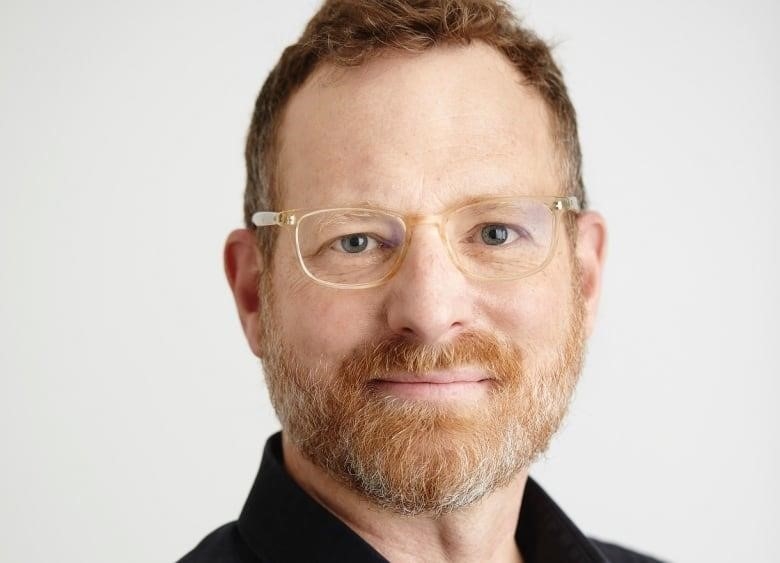 A man with red hair and clear glasses wears a black button-up shirt and poses for a photo.