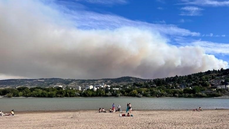 People in the distance along a long lakeshore watch a vast, long plume of white smoke billow over the lake from behind forested hills.
