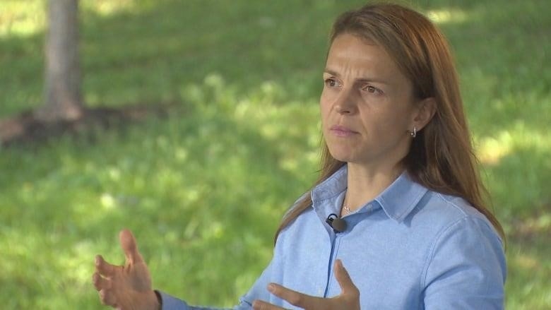 A woman standing with her hands out. She is wearing a light blue shirt and has long brown hair.