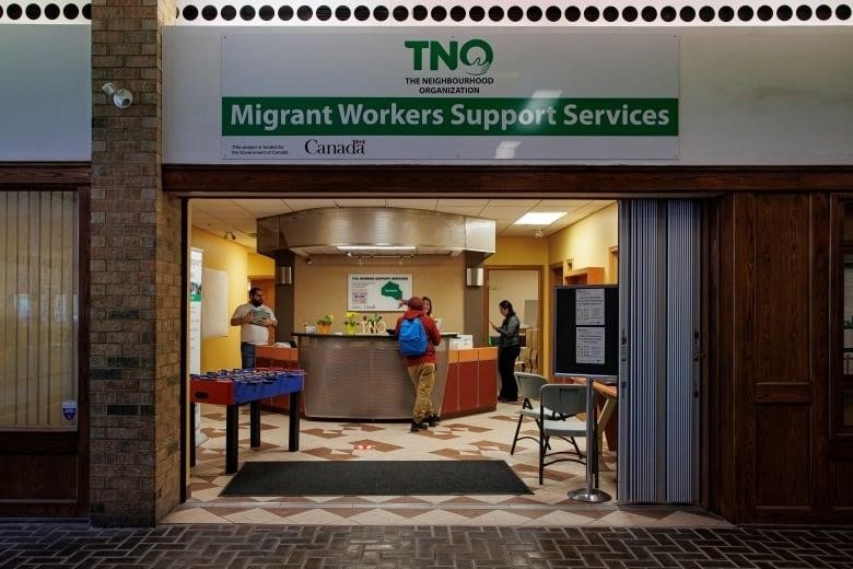 Three people stand near a counter with another person behind it, with a banner above the entrance that says: The Neighbourhood Organization; Migrant Workers Support Services.