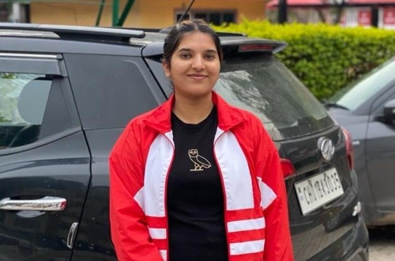 A woman wearing a red and white jacket and a black t-shirt stands in front of a black vehicle and smiles. 