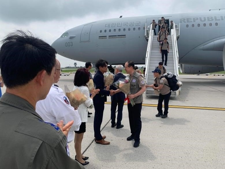 A line of people in uniform descend from an airplane and are greeted by people waiting on the tarmac.