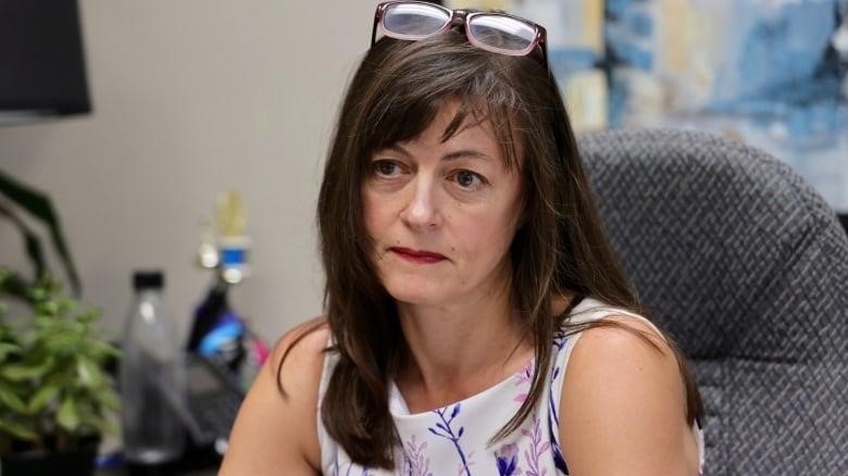 A white woman with long brown hair and glasses on her head sits behind a desk in an office. She has red lipstick and a white, floral dress