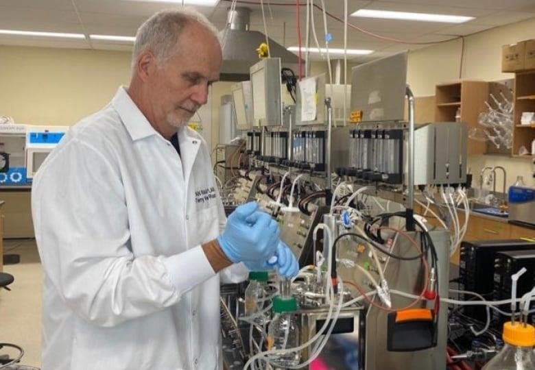 A man in a white lab coat and blue gloves works in a labratory in front of machines. 