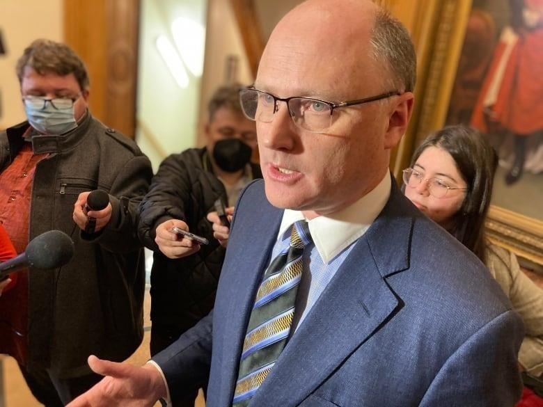 Bald man with glasses stands in lobby of legislature with reporters around him.