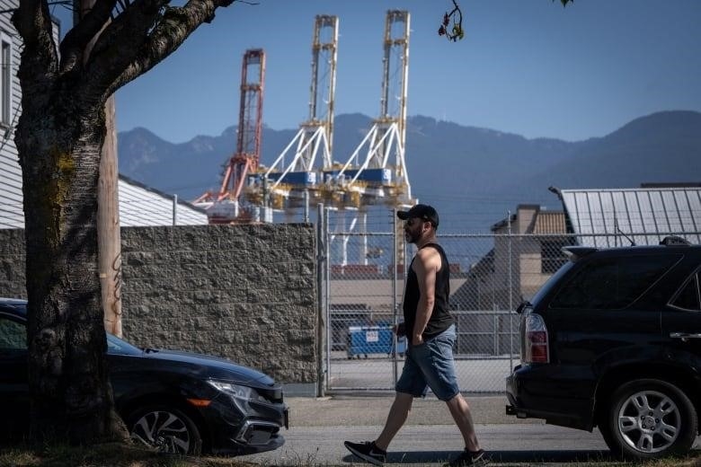 A man with a black tank top walks past a port with giant cargo cranes.