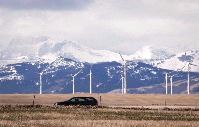An SUV drives along, with the backdrop of wind turbines in a field, and the snow-peaked Rockies behind.