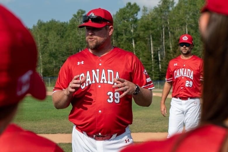 A man wearing a red Team Canada baseball jersey and hat speaks to players on a baseball field.