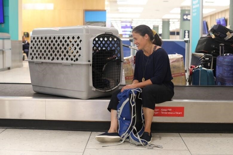 A woman sits by the conveyor belt at a baggage claims area with her dog in its crate.