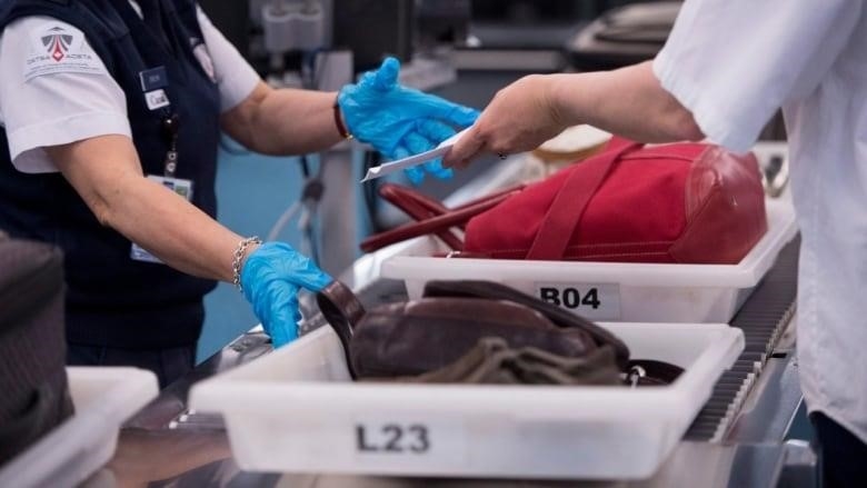 Security officer at an airport wears gloves and monitors travelers as they put their luggage in big plastic bins, which move down a conveyor belt.