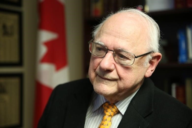 A man with thinning white hair and glasses stares at the camera. He's wearing a suit, a yellow tie and there's a Canada flag behind him.