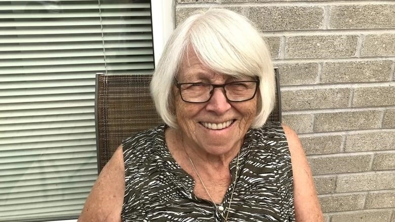 A woman with white hair sits in a chair and smiles.