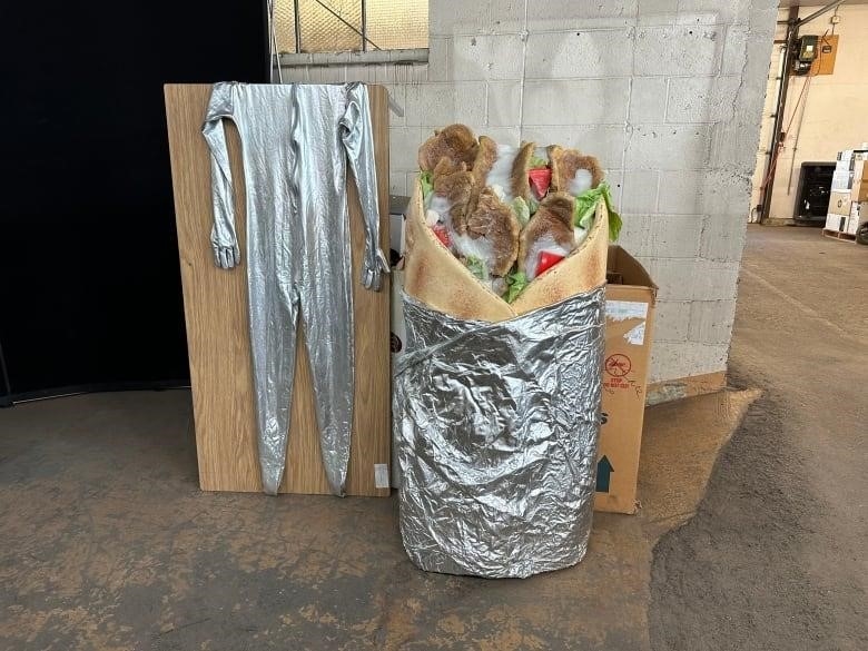 A person stands and waves dressed in donair costume, featuring tin foil, meat, white sauce, tomatoes and onion.