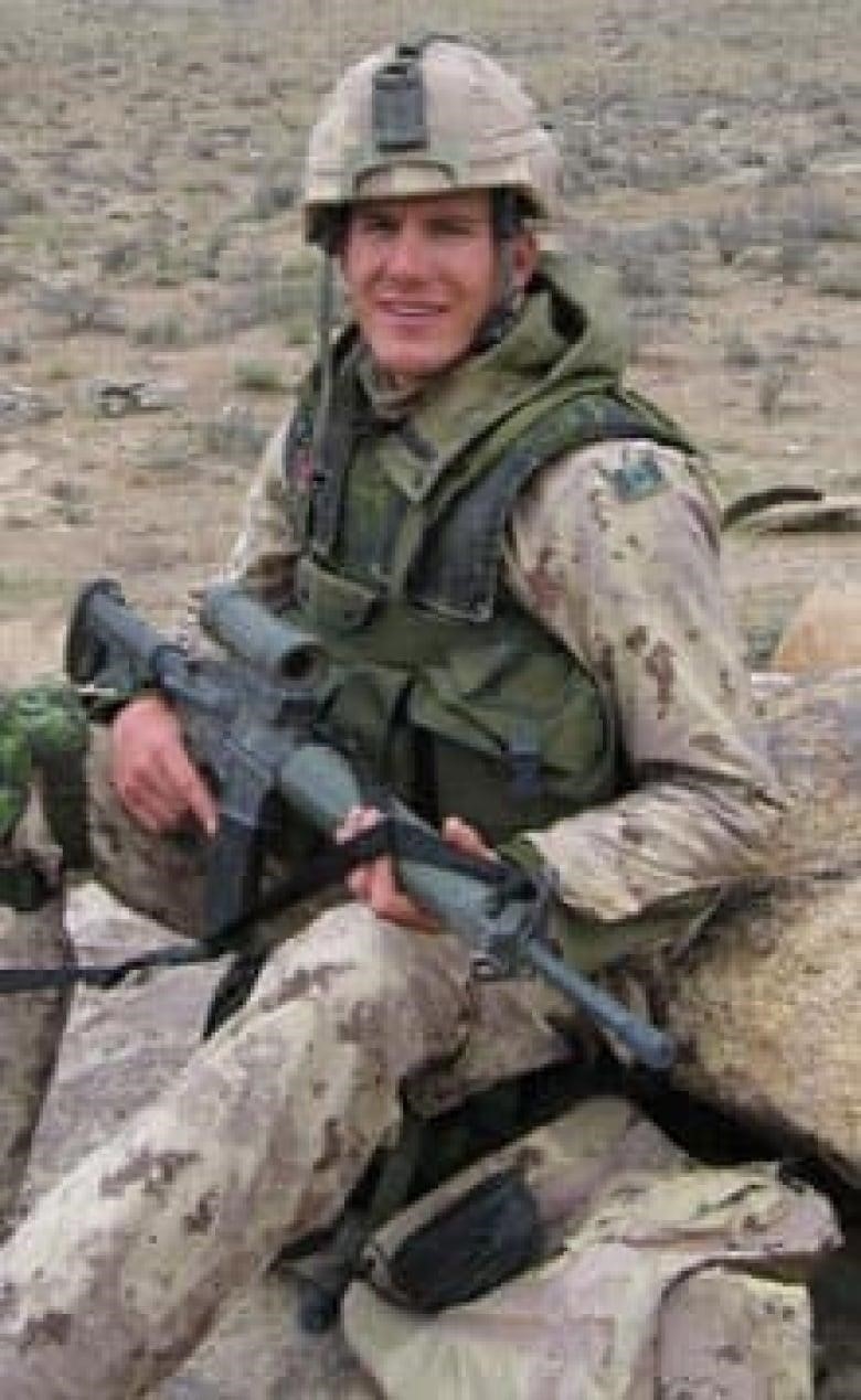 A man in a camouflage uniform sits with a rifle against some stones. 