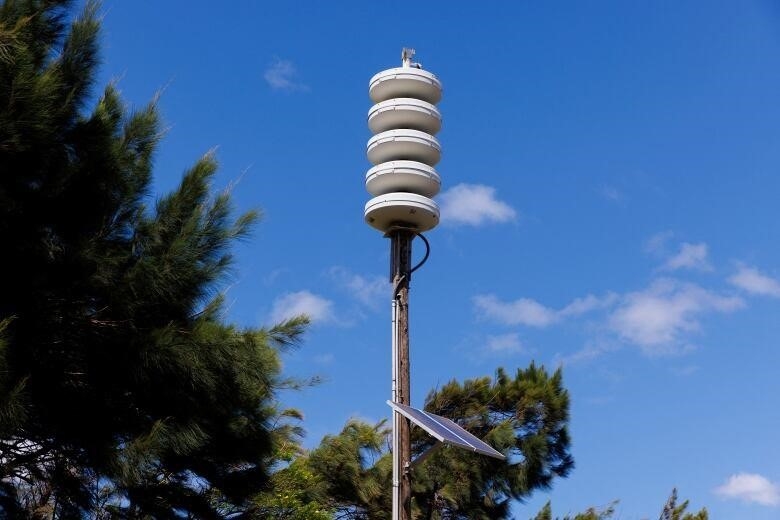 A cylindrical white siren is seen against a blue sky with trees in the background 