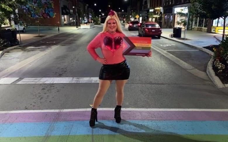 Drag queen holds up sign reading "everybody is welcome here" while standing on a rainbow crosswalk.