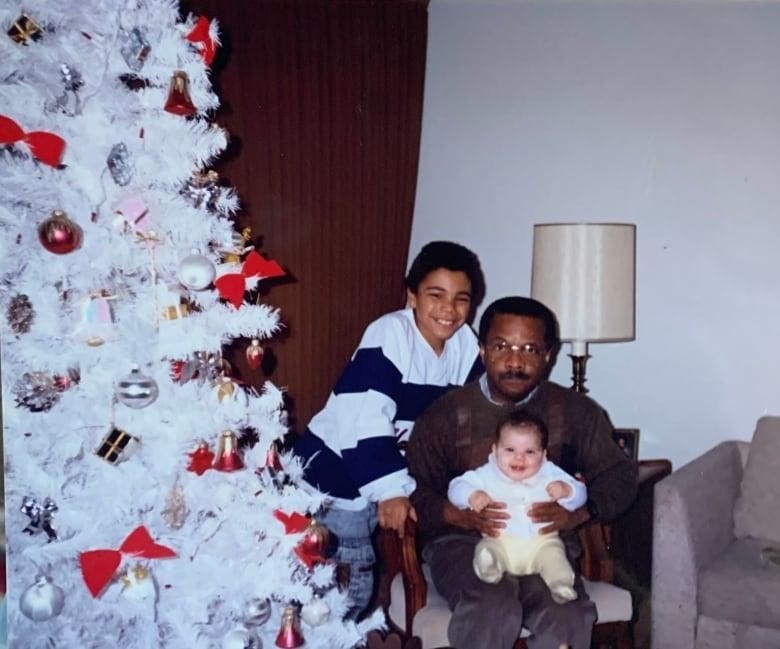 A reserved-looking man holds a smiling baby. They sit next to a white Christmas tree as a smiling boy leans next to them. 