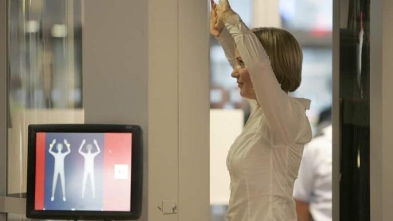 A woman in a white blouse stands with her arms above her head as she passes through a security scanner. To the left, a small screen shows a scan of her body. 