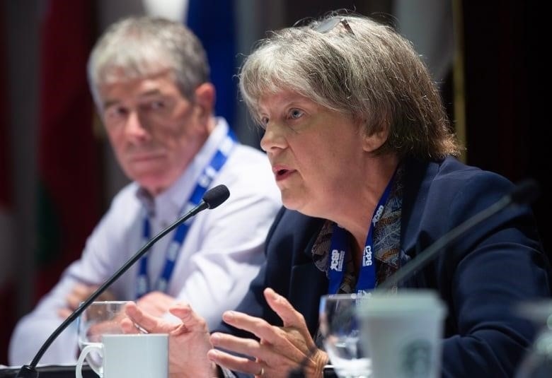 A greyhaired man looks at a woman while she is speaking at a podium. 