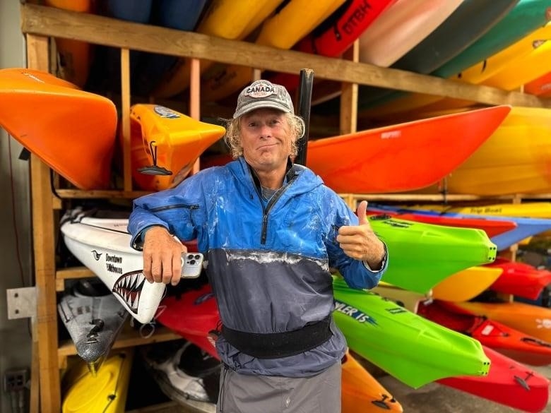A man wearing a blue coat and ballcap that says 'Canada' smiles at the camera and gives a thumbs up. He leans against a wall of colourful kayaks.
