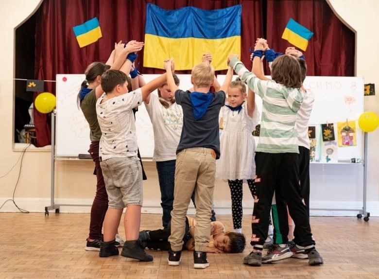 Children with their arms raised form a circle around a child lying on the floor. 