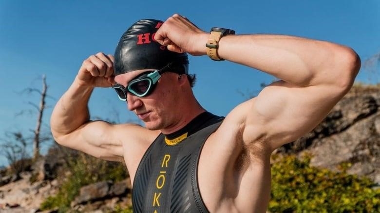 A man in a swimsuit and goggles flexes his sizeable arm muscles as he puts both hands on either side of his swim cap to adjust it.