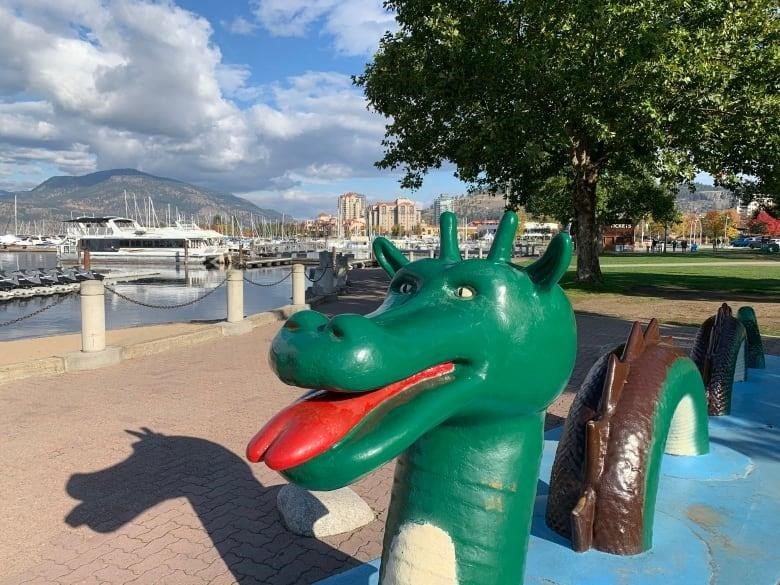 A green dragon-like statue is seen on the shore of a lake.