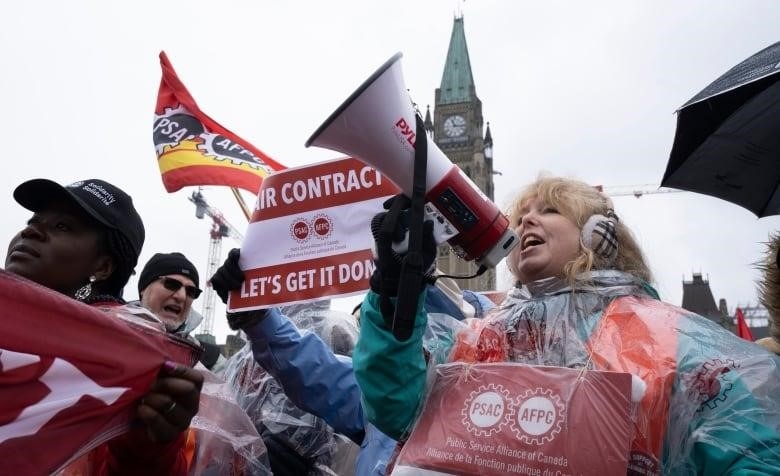 Striking government workers, including one with a megaphone, rally in front of a legislature on a drizzly spring day.