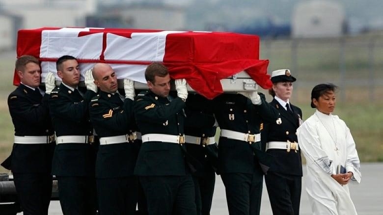 Several soldiers carry a casket draped in a Canadian flag in a procession led by a priest. 