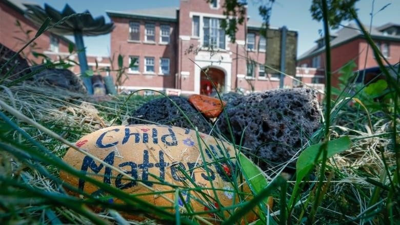 A hand painted stone that says "Every Child Matters" is seen amongst grass in the foreground, in front of a large red brick building.