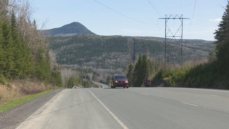 A vehicle and a motorcycle are pictured travelling in opposite directions on a highway. A mountain peak is visible in the background.