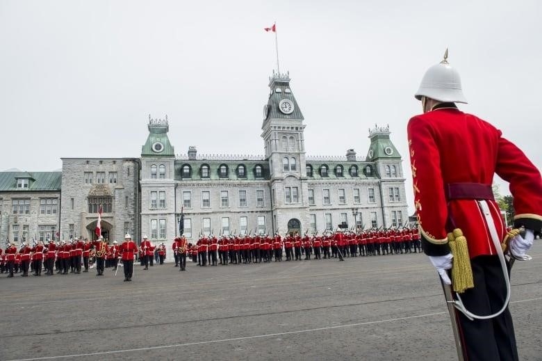 Officer cadets in red uniforms, black pants and white helmets are shown parading in front of a large, ornate brick building.