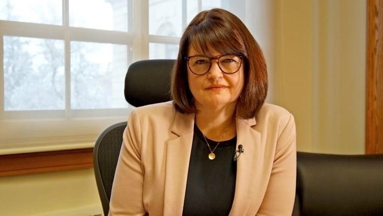 A woman with glasses sits in front of a window in an office.