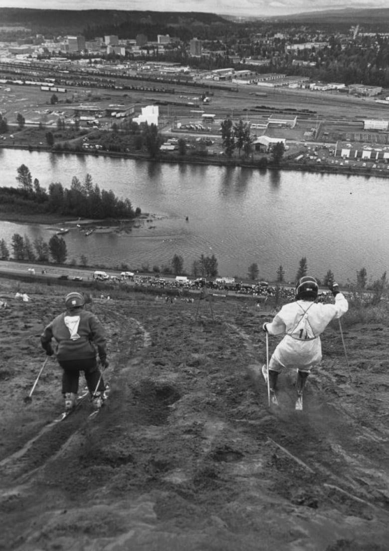 Two people on skis are shown from behind, skiing down a steep sandy slope, with a road, river and industrial area far down the hill in the distance.