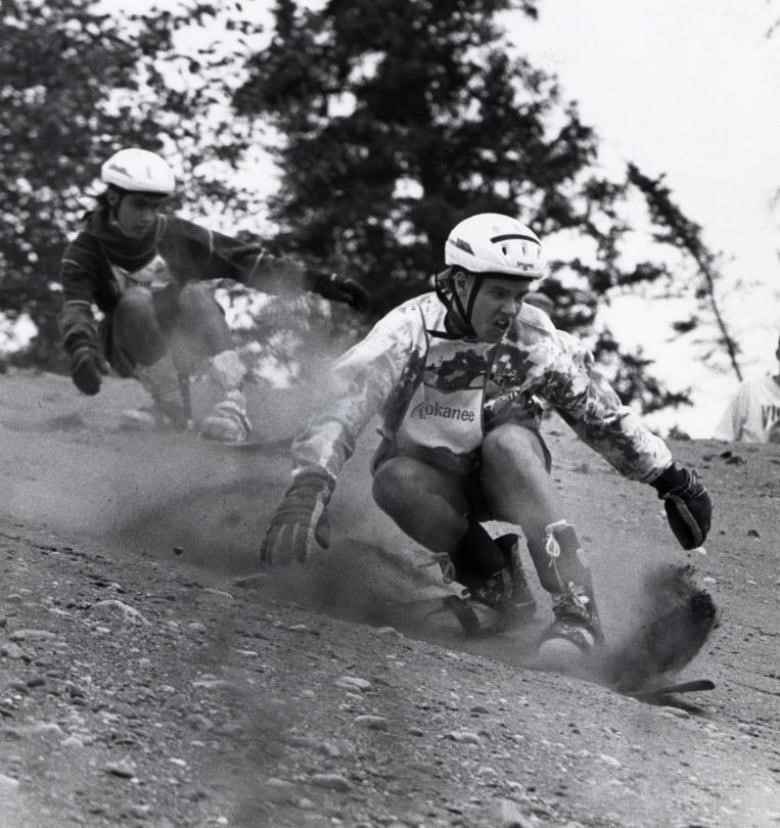 Two people ride snowboards down a sand and gravel slope, kicking up dust as they go.