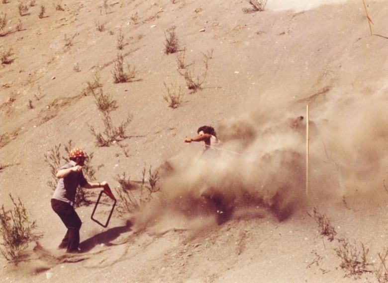 A person is shown losing control and kicking up a big cloud of dirt while trying to ski down a steep gravel slope.