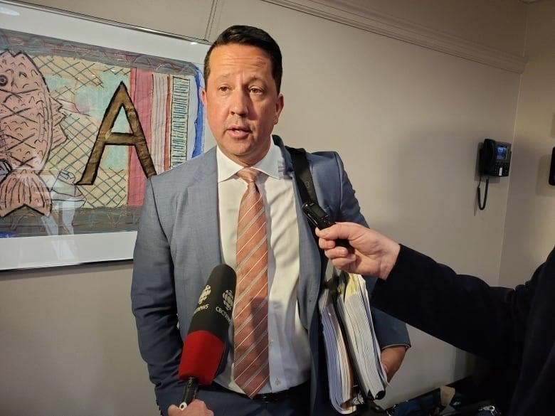 A man with short dark hair, wearing a grey blazer, white shirt and striped tie, holding a couple of binders full of papers under his left arm, speaks into reporters' microphones.