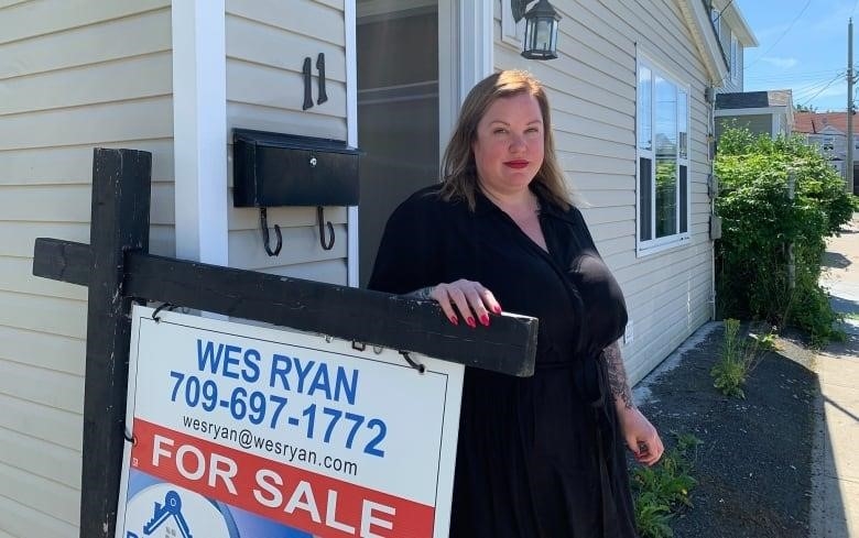 A woman wearing a black dress has her hand land on a for sale sign outside a beige home. 