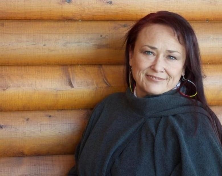 A woman in a black sweater stares ahead at the camera, in front of a building made of wood.