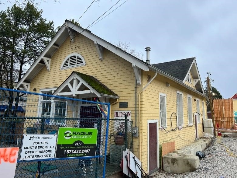 A small, one-storey yellow schoolhouse is surrounded by construction gates and signs.